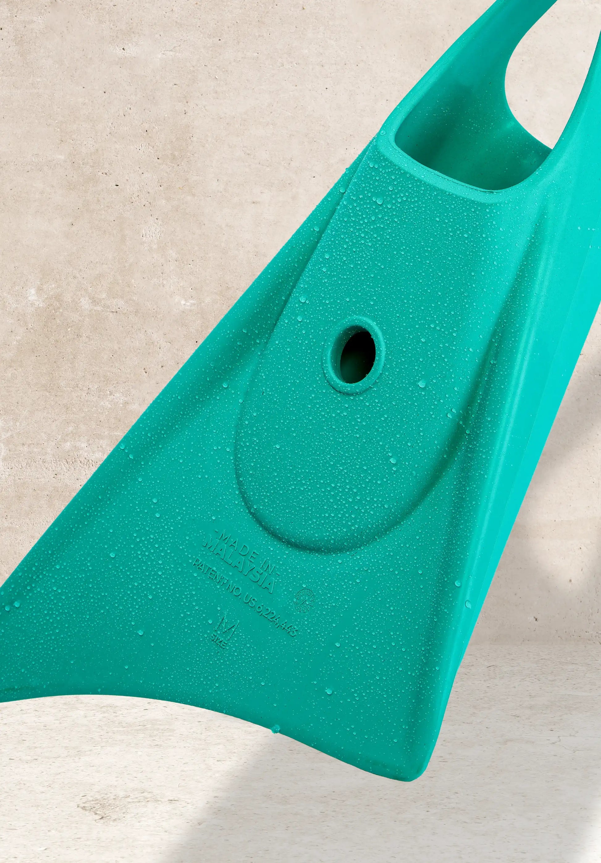 Teal bodyboard with water droplets, featuring handle and drain hole, resting on concrete surface.