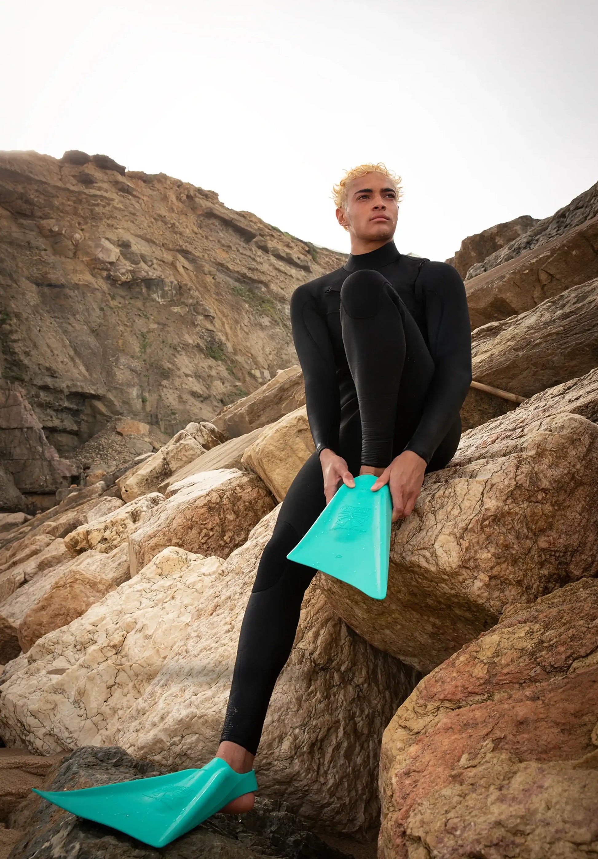 Surfer in black wetsuit holding teal boogie board on rocky coast, ready for wave riding