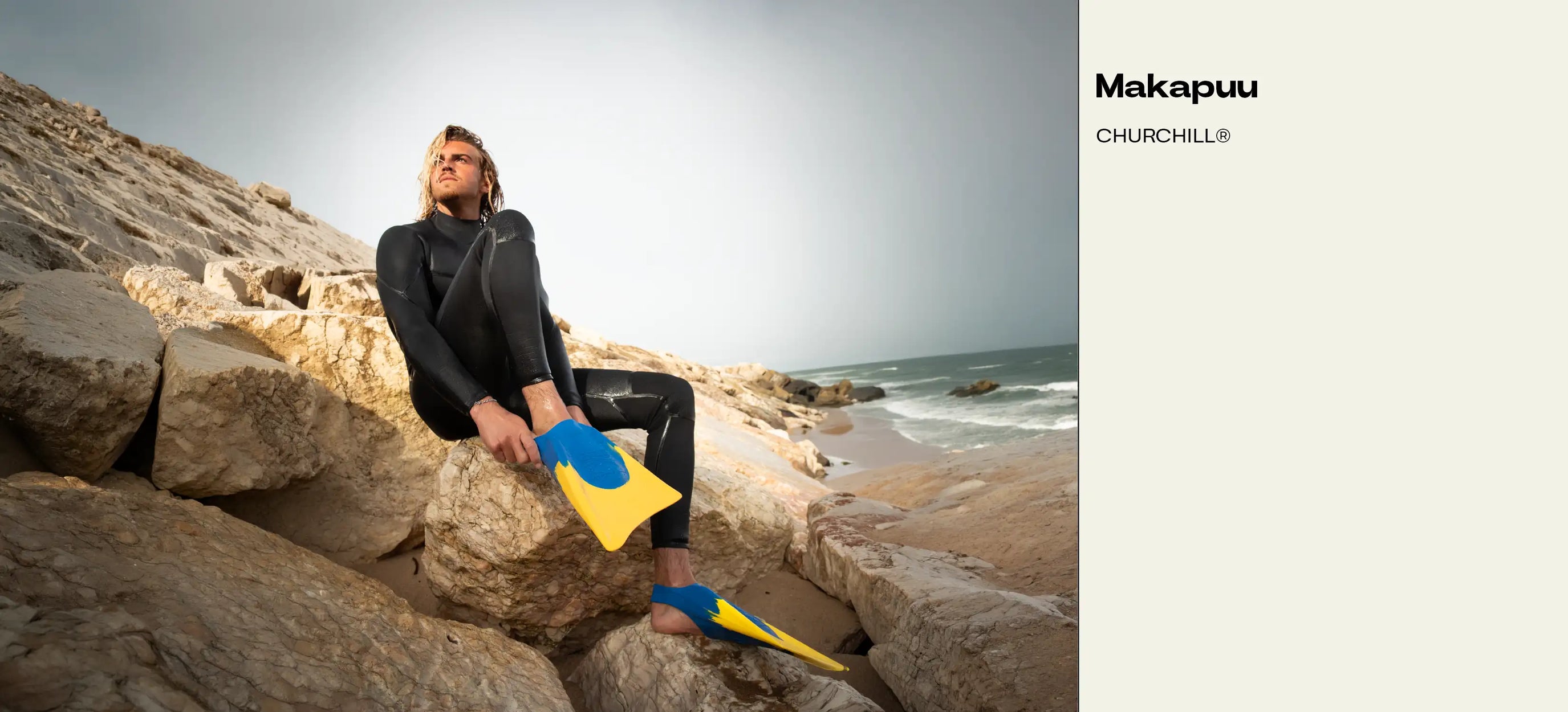 Man in wetsuit with blue and yellow fins sitting on coastal rocks near ocean waves