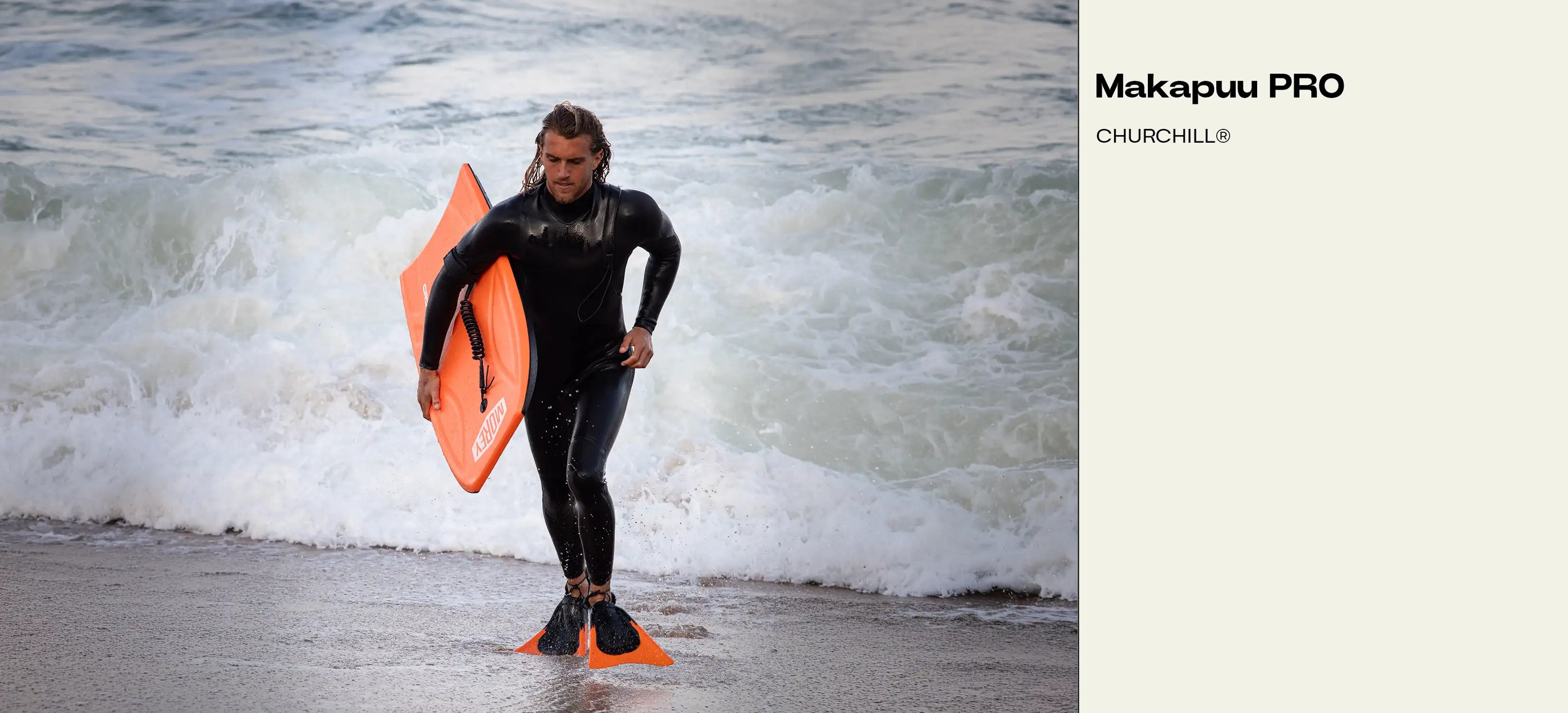 Man in wetsuit holding orange Makapuu PRO bodyboard at ocean shore with waves